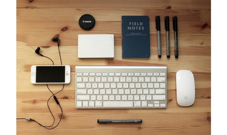 wooden table with modern tools such as a phone, earplugs, a keyboard, mouse and pencils laying on it. The image leaves a very organized impression.