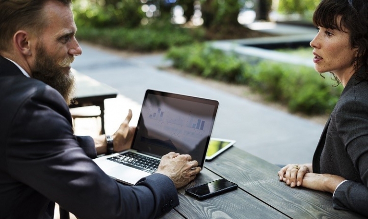 man with a beard in a suit sitting behind a laptop and talking to a women on the other side of the table.